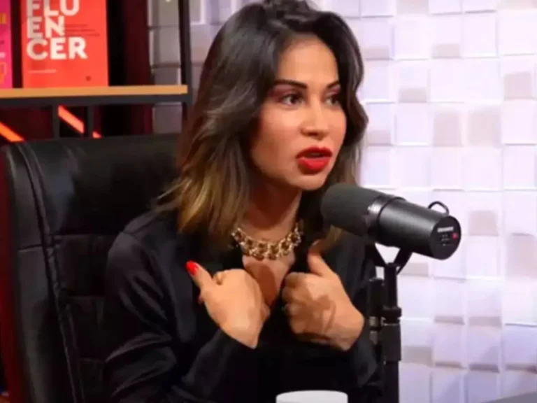 Maíra Cardi participa de um podcast, falando com gestos expressivos diante do microfone. Usando camisa preta, colar dourado e batom vermelho vibrante, a influenciadora aparece sentada em uma cadeira de couro, com fundo de estúdio decorado em tons claros e livros ao fundo. Sua postura firme reforça a intensidade do momento da entrevista.