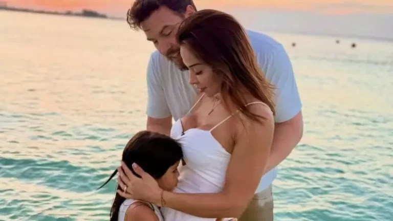 Maíra Cardi e Thiago Nigro posam em clima de ternura à beira-mar, durante o pôr do sol. De vestido branco, Maíra abraça a filha Sophia, enquanto Thiago, também de branco, envolve as duas com carinho. A cena transmite emoção e conexão familiar em um cenário paradisíaco.