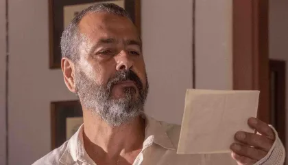 Marcos Palmeira surge em cena contemplativa, segurando uma carta ou fotografia com olhar atento. Com barba grisalha e camisa clara, o ator transmite seriedade e introspecção em um ambiente interno de decoração simples. A imagem reforça a densidade emocional do momento, possivelmente ligado a uma trama dramática.