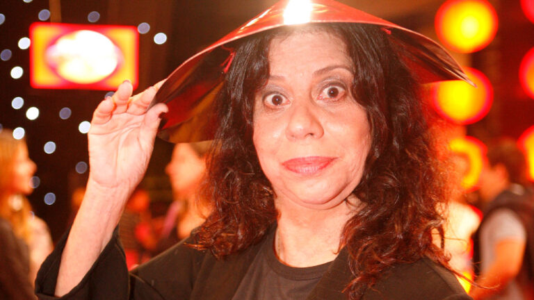 Maria Gladys segura adereço metálico na cabeça durante evento com iluminação vermelha ao fundo