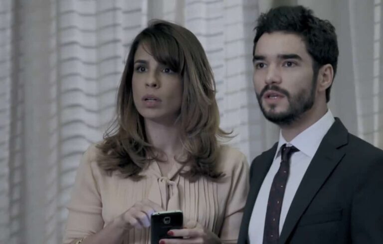 Maria Ribeiro e Caio Blat aparecem em cena da novela Império (2014). Os atores surgem lado a lado, com expressões de surpresa e preocupação. Maria segura um celular enquanto usa uma blusa clara, e Caio veste terno escuro e gravata, em um ambiente de decoração sofisticada, típico da trama assinada por Aguinaldo Silva.