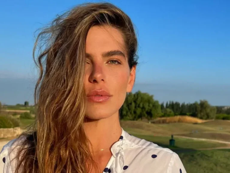 Mariana Goldfarb posa ao ar livre em um cenário de campo aberto sob céu azul intenso. A modelo exibe expressão serena, com os cabelos castanhos soltos e ondulados. Ela veste uma blusa branca com estampa de bolinhas, em um clique que valoriza a luz natural e o clima leve e bucólico do ambiente.