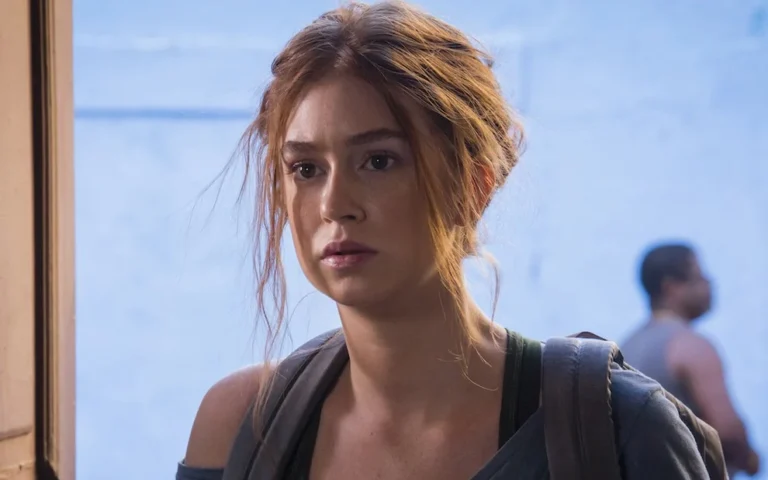Marina Ruy Barbosa interpreta Eliza na novela Totalmente Demais. Na cena, a personagem aparece com expressão séria e olhar pensativo, usando uma blusa cinza e mochila nos ombros. Com os cabelos ruivos presos de forma despojada e alguns fios soltos, Eliza transmite a determinação e a vulnerabilidade características de sua trajetória na trama.