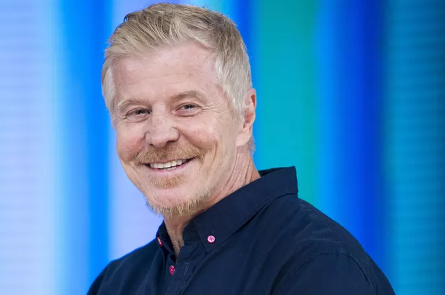 Miguel Falabella sorri para a câmera em ambiente de estúdio, vestindo camisa azul marinho. Com visual elegante e cabelo claro, o ator, diretor e autor transmite simpatia em frente a um fundo colorido em tons de azul e verde
