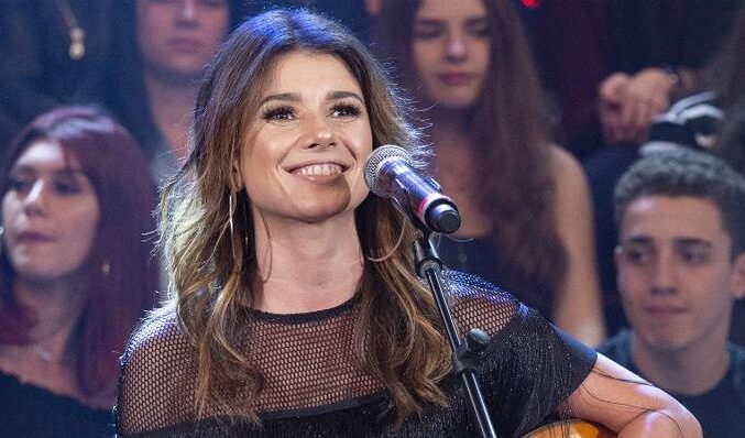Paula Fernandes sorri durante uma apresentação musical ao vivo. A cantora, usando uma blusa preta com transparência e brincos de argola, segura o violão e se posiciona diante do microfone. Ao fundo, a plateia acompanha atenta o momento, reforçando o clima intimista e animado do show.