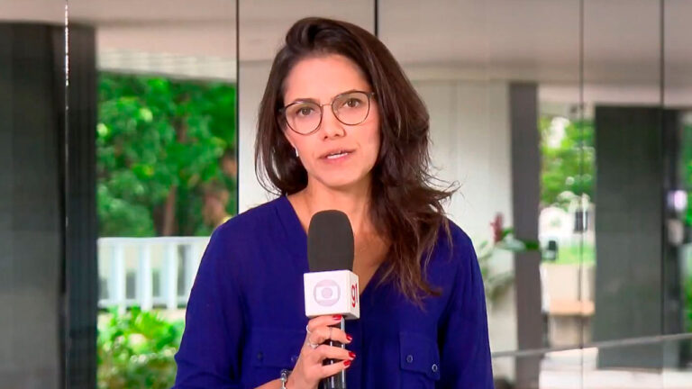 Raquel Honorato, ex-repórter do Jornal Hoje, segura microfone da Globo durante reportagem em área externa com vidros e vegetação ao fundo