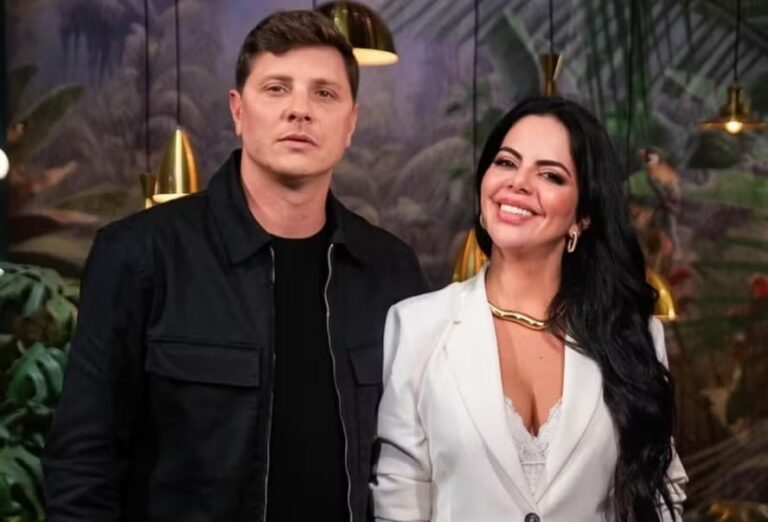 Renata Giaffredo e Alexandre Thomaz posam juntos em um ambiente decorado com plantas e luminárias douradas. Renata sorri para a câmera, vestindo um blazer branco elegante e acessórios dourados, enquanto Alexandre, com expressão séria, usa uma jaqueta preta sobre camiseta escura. O cenário transmite um clima sofisticado e descontraído.