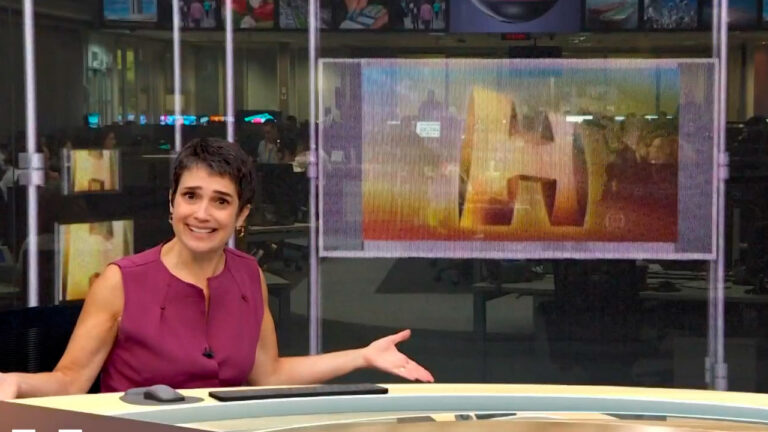 Sandra Annenberg apresenta o Jornal Hoje sorrindo em frente a telão com logotipo do telejornal