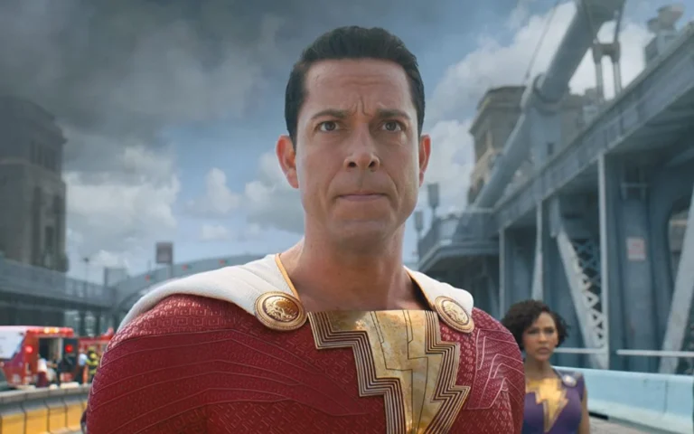 Cena do filme Shazam!, com Zachary Levi caracterizado como o herói Shazam. Usando o icônico traje vermelho com símbolo dourado no peito e capa branca, o personagem aparece com expressão séria em uma ponte, sob um céu nublado. Ao fundo, uma heroína uniformizada observa a situação, reforçando o clima de tensão e ação do momento.