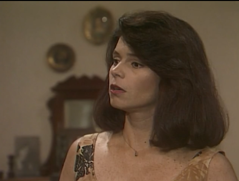 Françoise Forton aparece em cena da novela Tieta (1989). Com expressão atenta, a atriz usa um vestido claro com detalhes escuros e um colar delicado. Seu cabelo volumoso e bem arrumado completa o visual elegante da personagem, em um ambiente interno decorado com espelhos e móveis clássicos ao fundo.