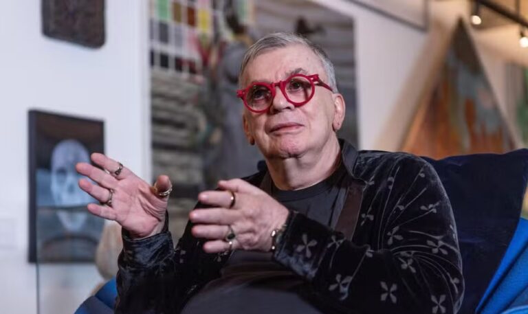 Walcyr Carrasco aparece sentado em um ambiente artístico, rodeado por quadros e obras de arte. O autor usa óculos vermelhos chamativos, anéis nas mãos e veste uma blusa escura com detalhes de estampa discreta. Com expressão atenta e gesticulando enquanto fala, ele transmite concentração e entusiasmo durante a conversa.