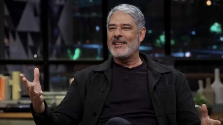 William Bonner aparece descontraído durante entrevista no Conversa com Bial. De barba grisalha e usando roupas escuras, o jornalista sorri enquanto gesticula com as mãos, em um cenário de estúdio moderno com luzes ao fundo