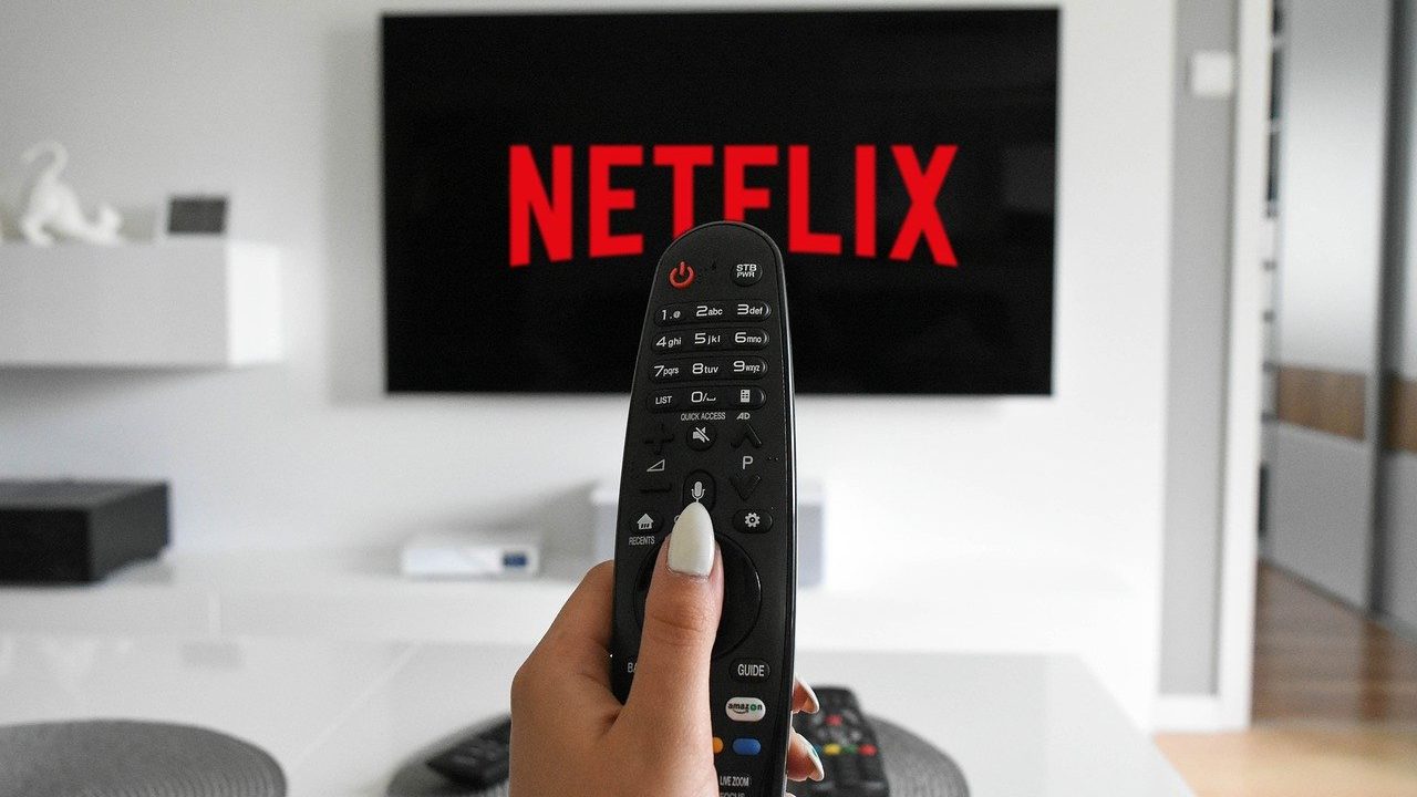 Imagem com foto de uma mão feminina segurando o controle remoto com o logo da Netflix ao fundo na televisão