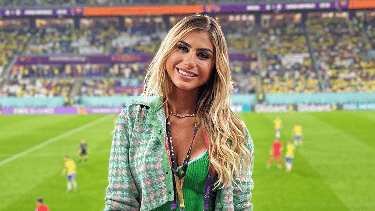 Imagem com foto da jornalista Renata Heilborn dentro de um estádio de futebol. A repórter está sorrindo e ao fundo, no gramado do campo, estão jogadores durante uma partida.