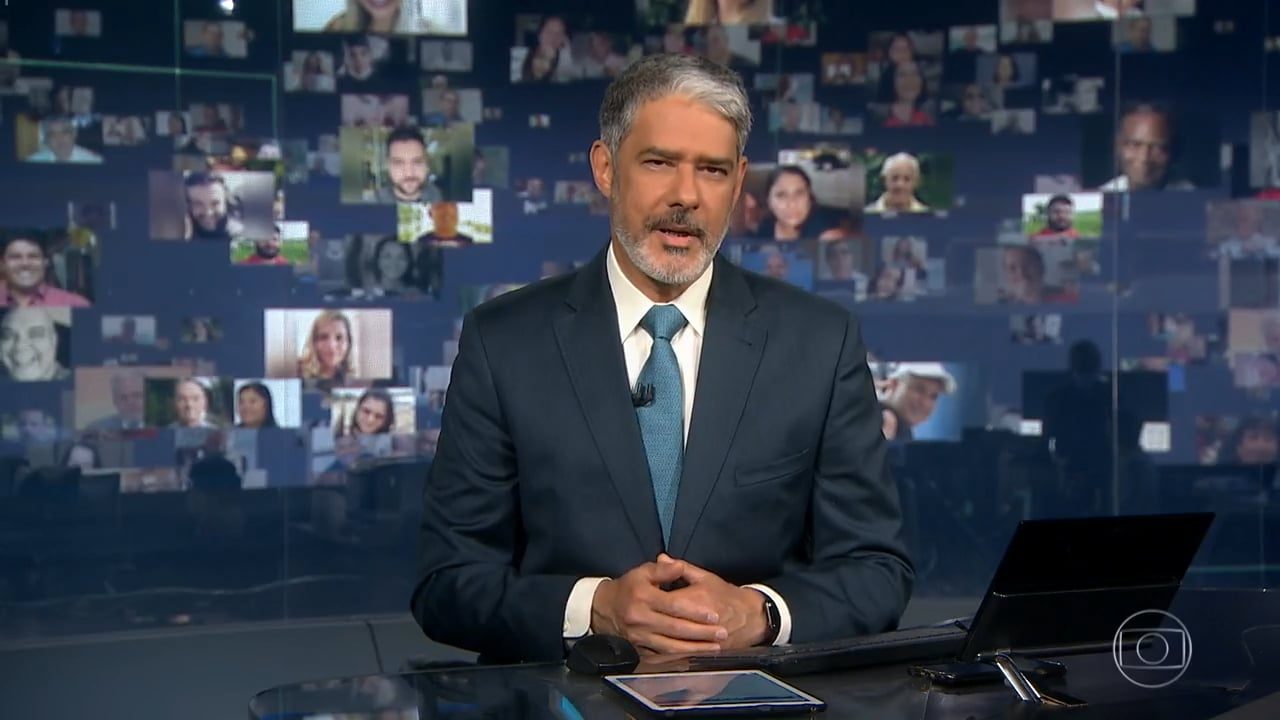 Imagem do apresentador William Bonner no Jornal Nacional com fotos das vítimas da crise sanitária projetadas ao fundo