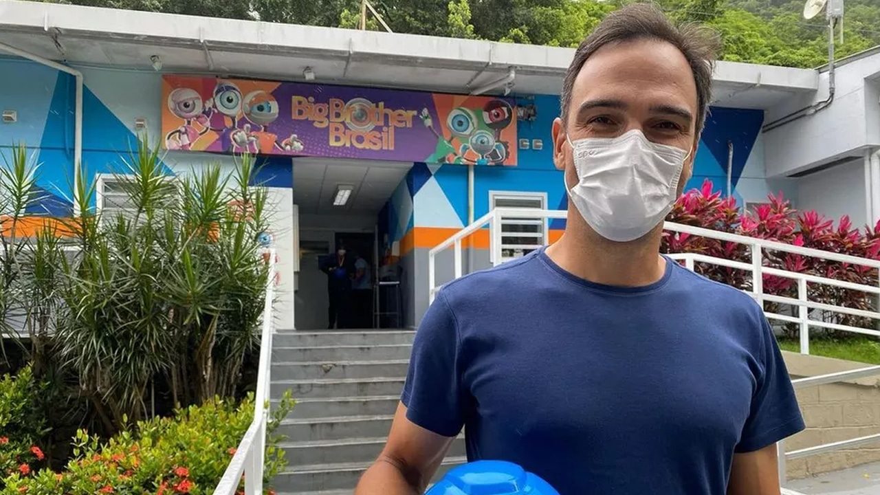 Imagem com foto do apresentador Tadeu Schmidt na porta dos estúdios do Big Brother Brasil. Ele está de máscara de proteção e segura um capacete da cor azul com um adesivo do logo do programa. O texto fala sobre a divulgação dos participantes do BBB