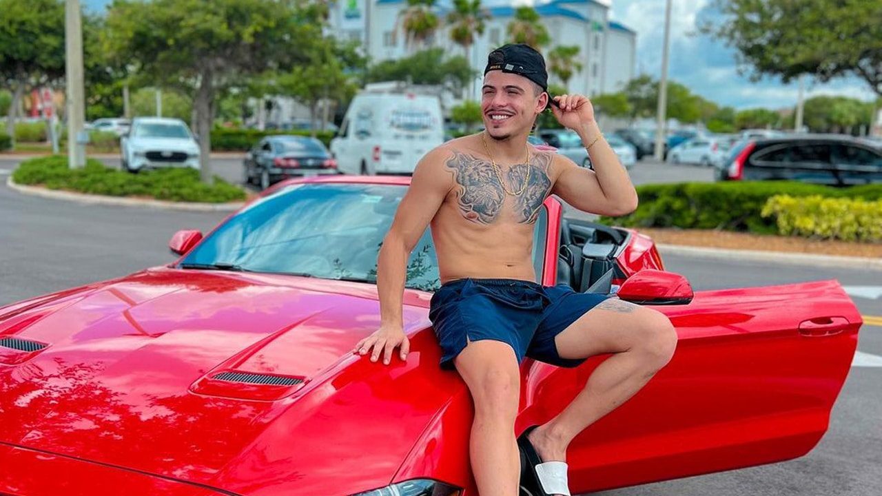 Imagem com foto do ator Thomaz Costa sentado em um carro de luxo vermelho