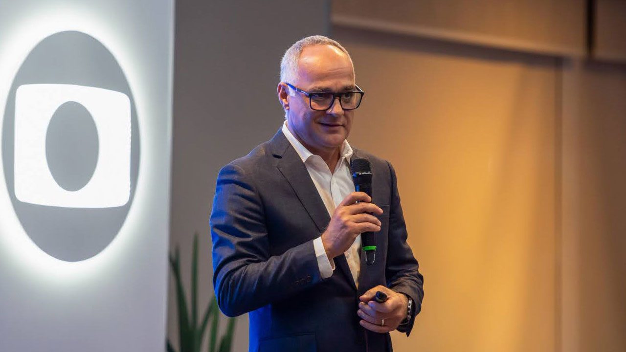 Amauri Soares, executivo da TV Globo, segurando um microfone durante apresentação, com o logotipo da emissora projetado ao fundo