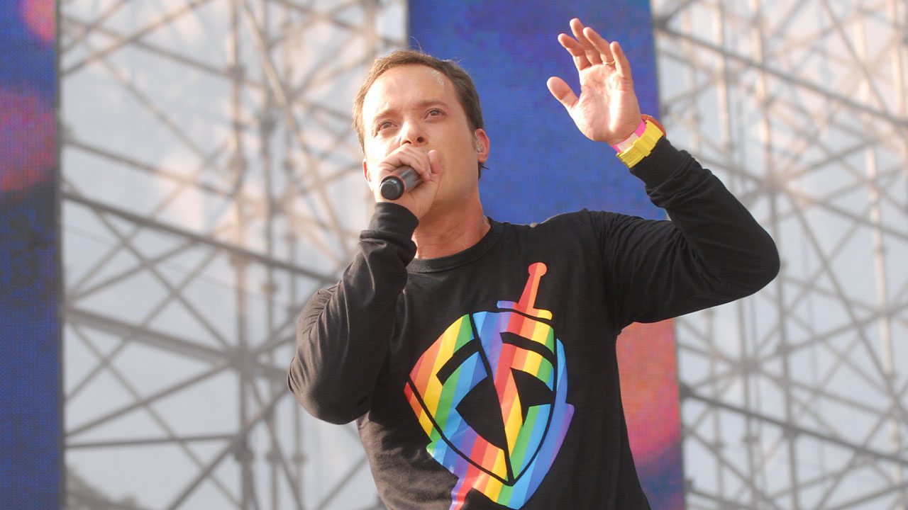 André Valadão cantando no palco com microfone na mão e camiseta com símbolo colorido em evento gospel