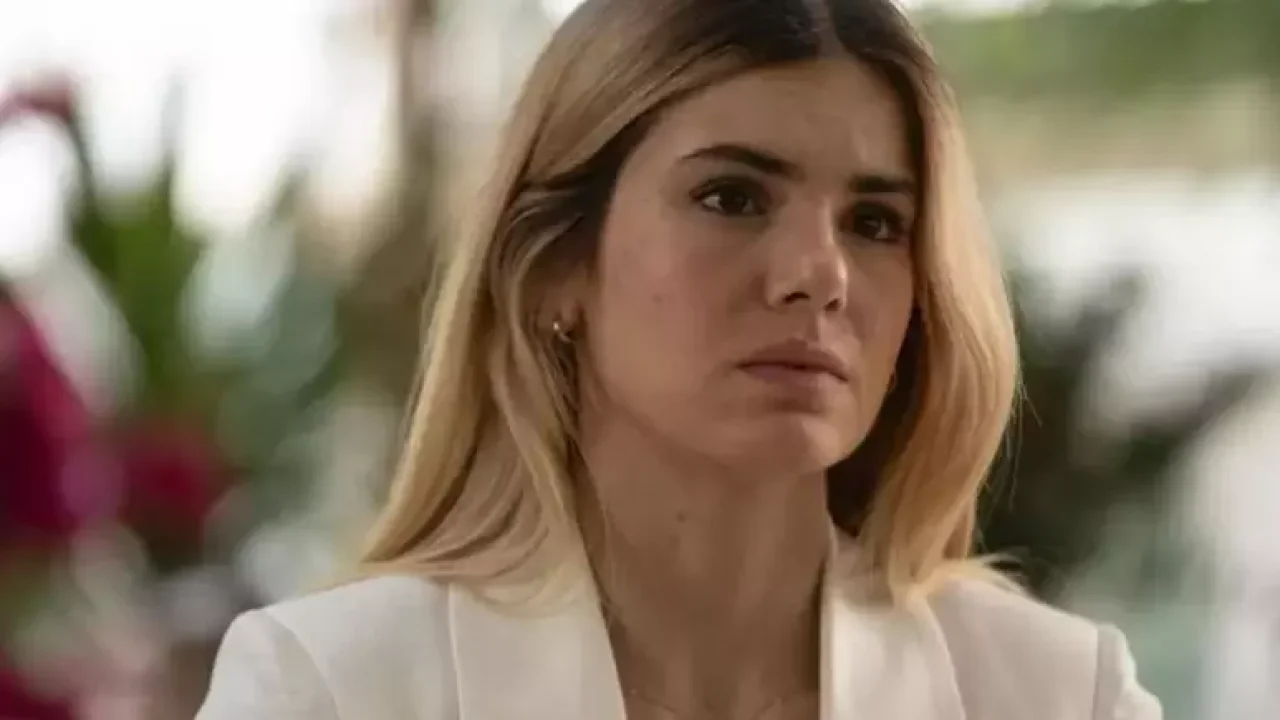 A imagem mostra a atriz Camila Queiroz em uma cena da novela Beleza Fatal. Ela aparece com uma expressão séria e intensa, vestindo um blazer branco, o que transmite um ar de sofisticação e poder. Seu cabelo loiro está solto e levemente ondulado, e o fundo desfocado sugere um ambiente elegante, possivelmente uma cena de grande carga dramática