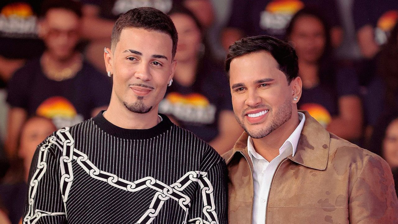 Carlinhos Maia e Lucas Guimarães no palco do Teleton, sorrindo enquanto posam lado a lado, com plateia ao fundo vestindo camisetas da campanha