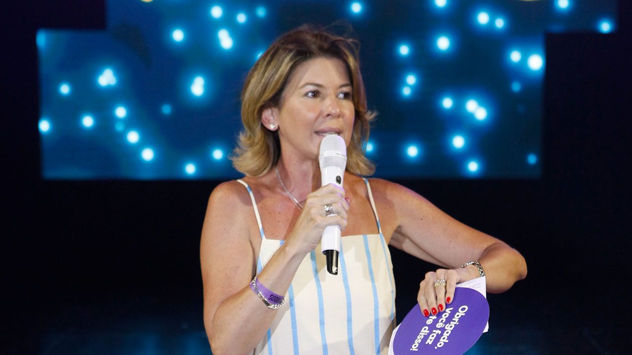 Daniela Beyruti segurando microfone e folder roxo durante evento do SBT com painel iluminado ao fundo