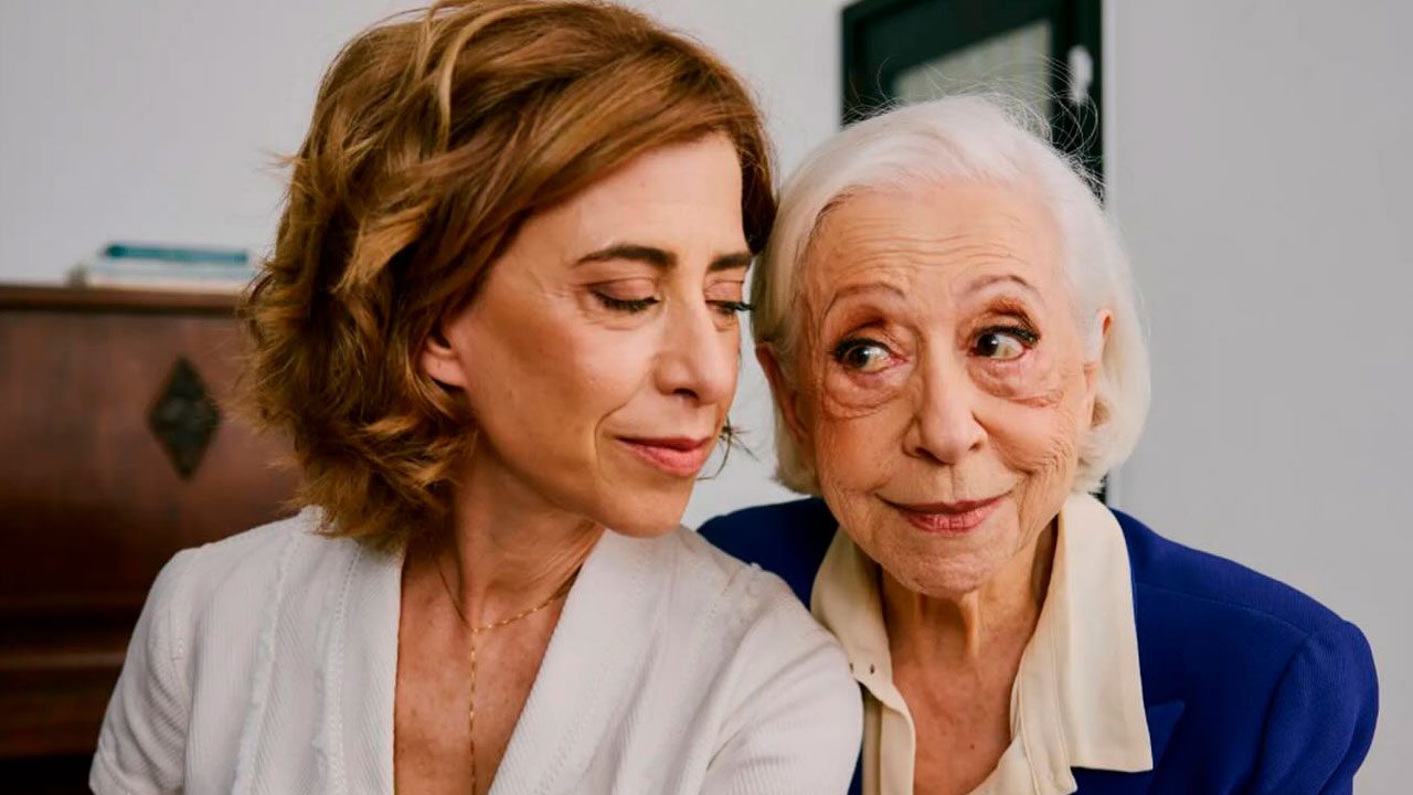 Fernanda Torres e Fernanda Montenegro sentadas lado a lado, trocando olhares carinhosos, com um piano ao fundo em ambiente aconchegante