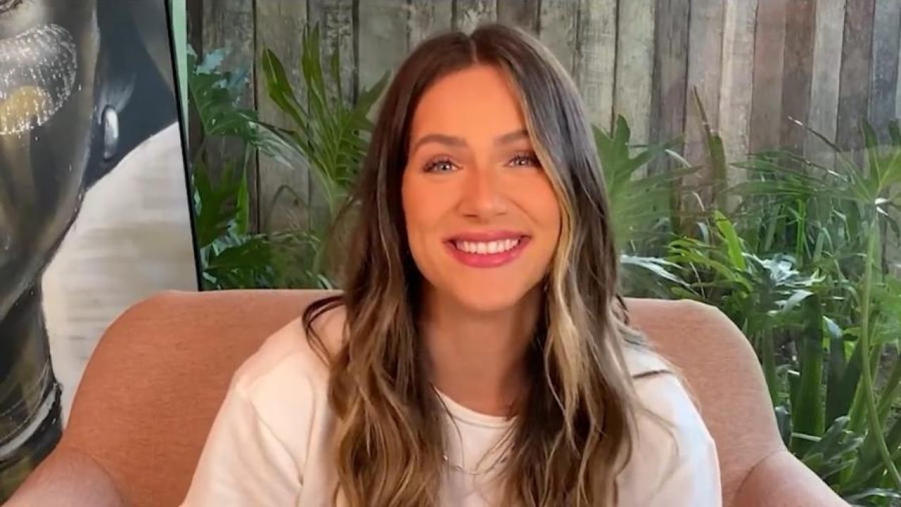 Giovanna Ewbank sorri para a câmera durante gravação em ambiente descontraído. Usando camiseta branca, a apresentadora aparece sentada em uma poltrona, com os cabelos soltos e levemente ondulados, diante de um fundo com plantas e uma obra de arte ao lado