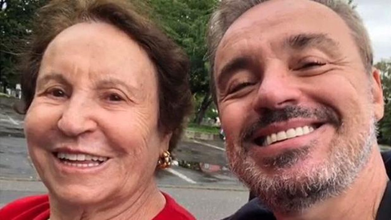 A imagem mostra o apresentador Gugu Liberato ao lado de sua mãe, Maria do Céu. Os dois aparecem sorrindo em um ambiente ao ar livre, transmitindo um momento de carinho e cumplicidade