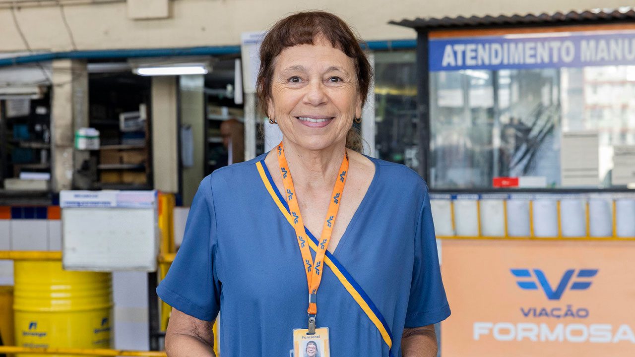 Iara Jamra sorrindo enquanto usa uniforme azul e crachá da Viação Formosa em um ambiente de atendimento da empresa fictícia da novela Volta por Cima