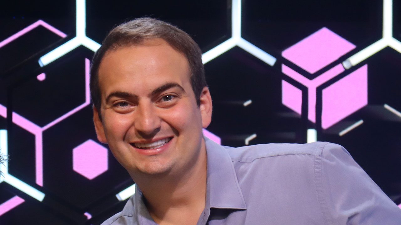 João Paulo Cappellanes, comentarista da Band, sorrindo em cenário com luzes geométricas