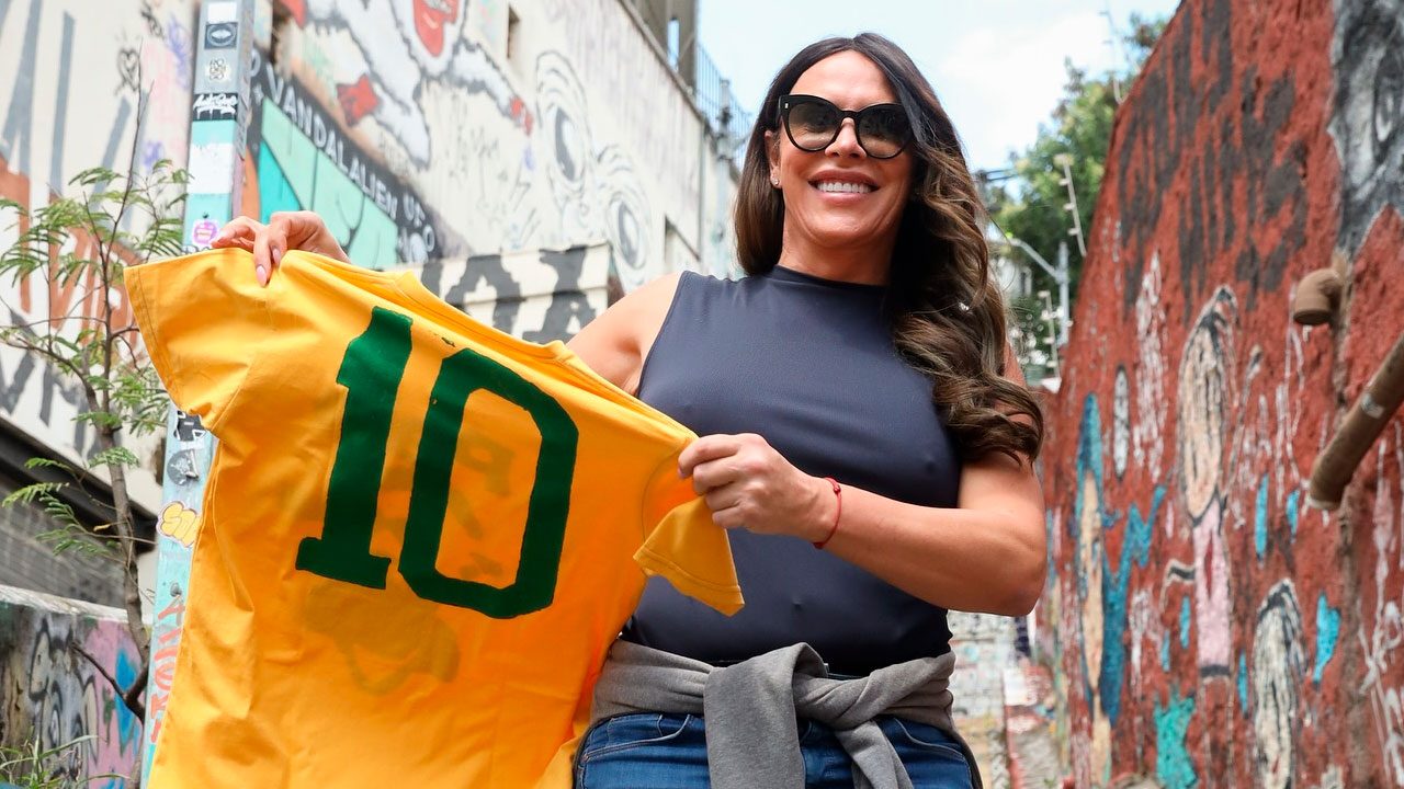 Karla Sofía Gascón sorri enquanto segura uma camisa amarela com o número 10 estampado, em um local com paredes grafitadas ao fundo