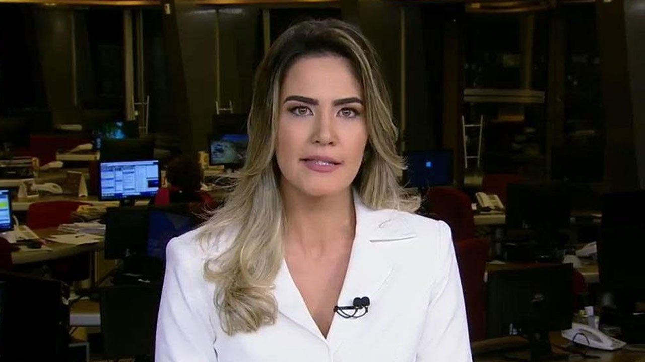 Larissa Alvarenga apresenta o telejornal Fala Brasil, vestindo blazer branco e microfone de lapela, com a redação da emissora ao fundo