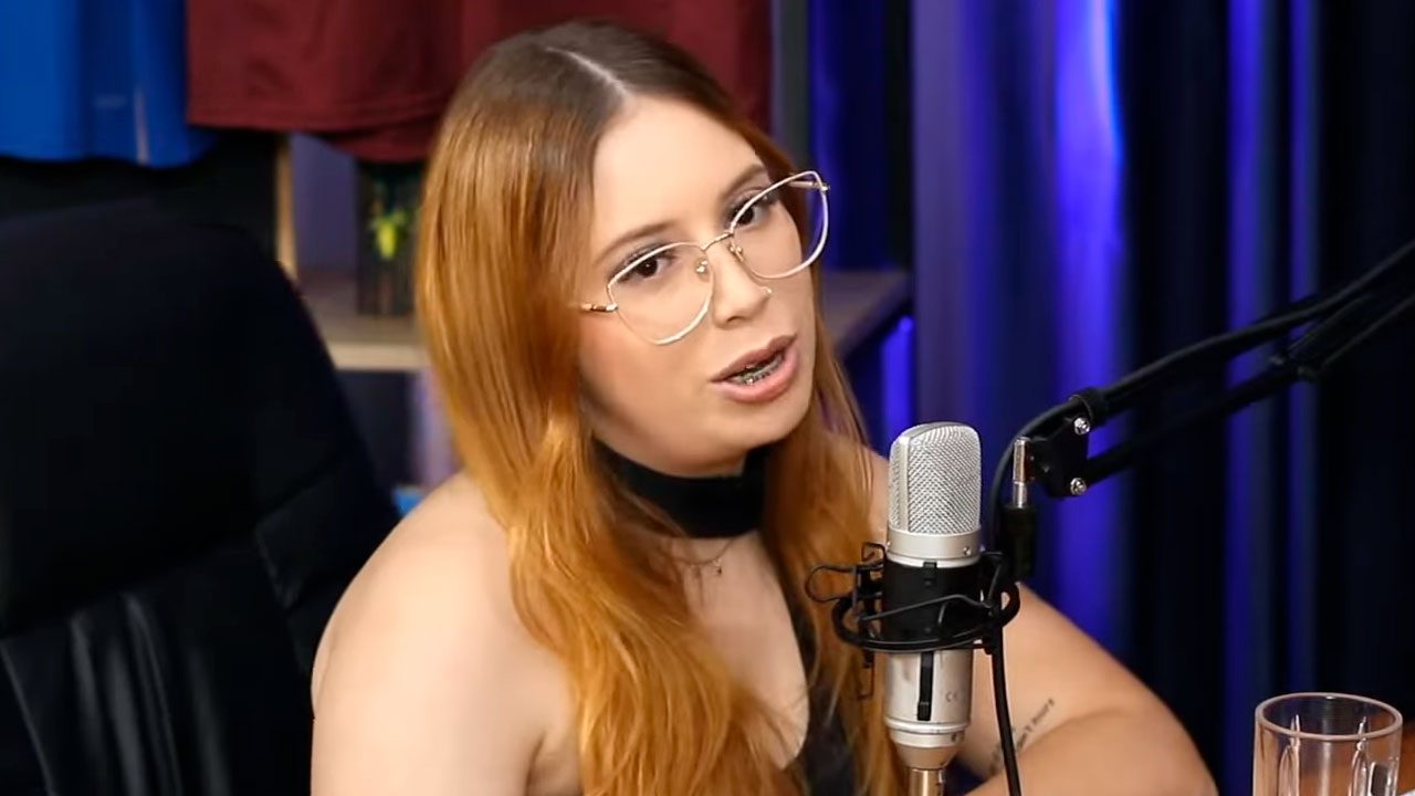 Influenciadora Luana Santos durante participação em um podcast, falando ao microfone com fundo escuro iluminado em azul, usando óculos e cabelo ruivo solto