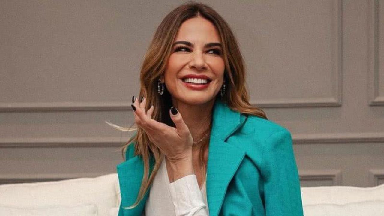 Na imagem, vemos Luciana Gimenez em um momento descontraído e sorridente. Ela está sentada em um ambiente sofisticado, com um fundo cinza de painéis elegantes. Seu olhar e expressão transmitem alegria e leveza