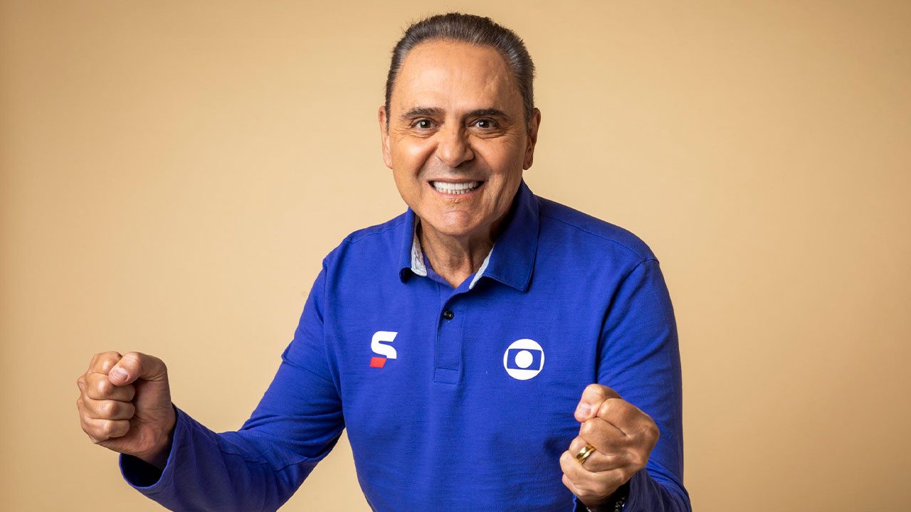 Luis Roberto sorrindo para a câmera, vestindo camisa azul com o logotipo do SporTV e da Globo, fazendo gesto de entusiasmo com os punhos cerrados