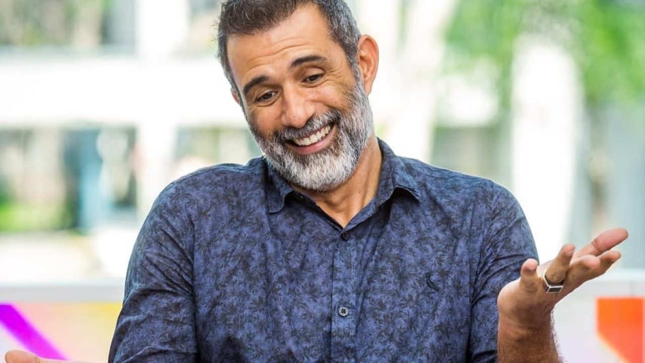 Marcos Pasquim aparece sorridente e descontraído durante entrevista, com os braços abertos em gesto leve de bom humor. O ator usa camisa azul estampada e exibe visual com barba grisalha, em cenário iluminado e colorido ao fundo.