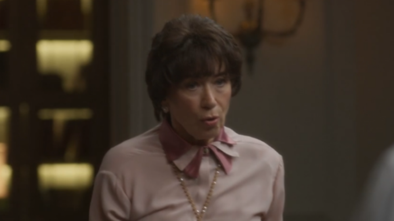 Cena da novela Garota do Momento, da TV Globo, com a personagem Maristela, interpretada por Lilia Cabral. Na imagem, ela aparece com expressão séria, vestindo uma camisa rosa de laço e um colar dourado, em um ambiente interno com iluminação suave e decoração clássica ao fundo