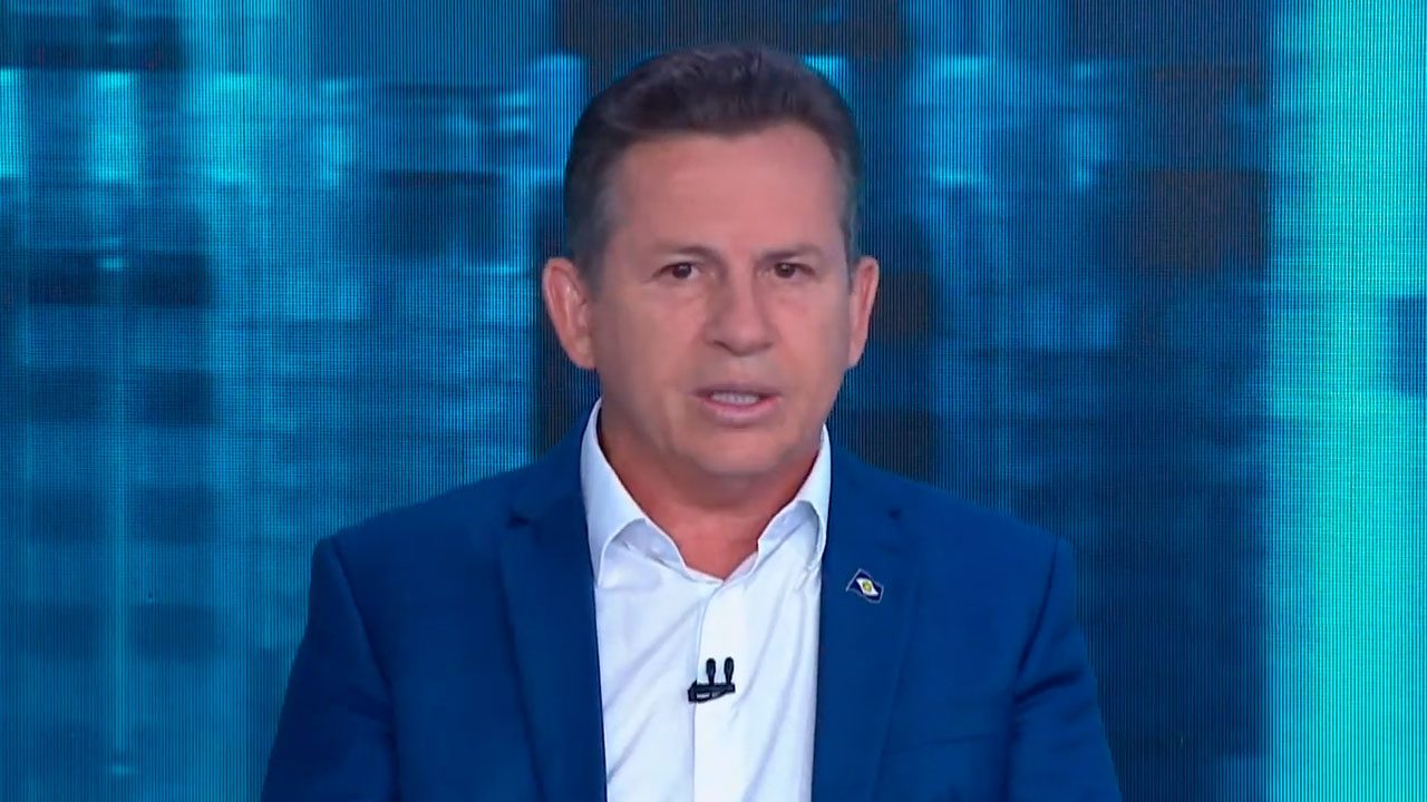 Mauro Mendes, governador do Mato Grosso, durante entrevista no programa Direto ao Ponto, da Jovem Pan, usando terno azul e falando diante de um fundo digital azul