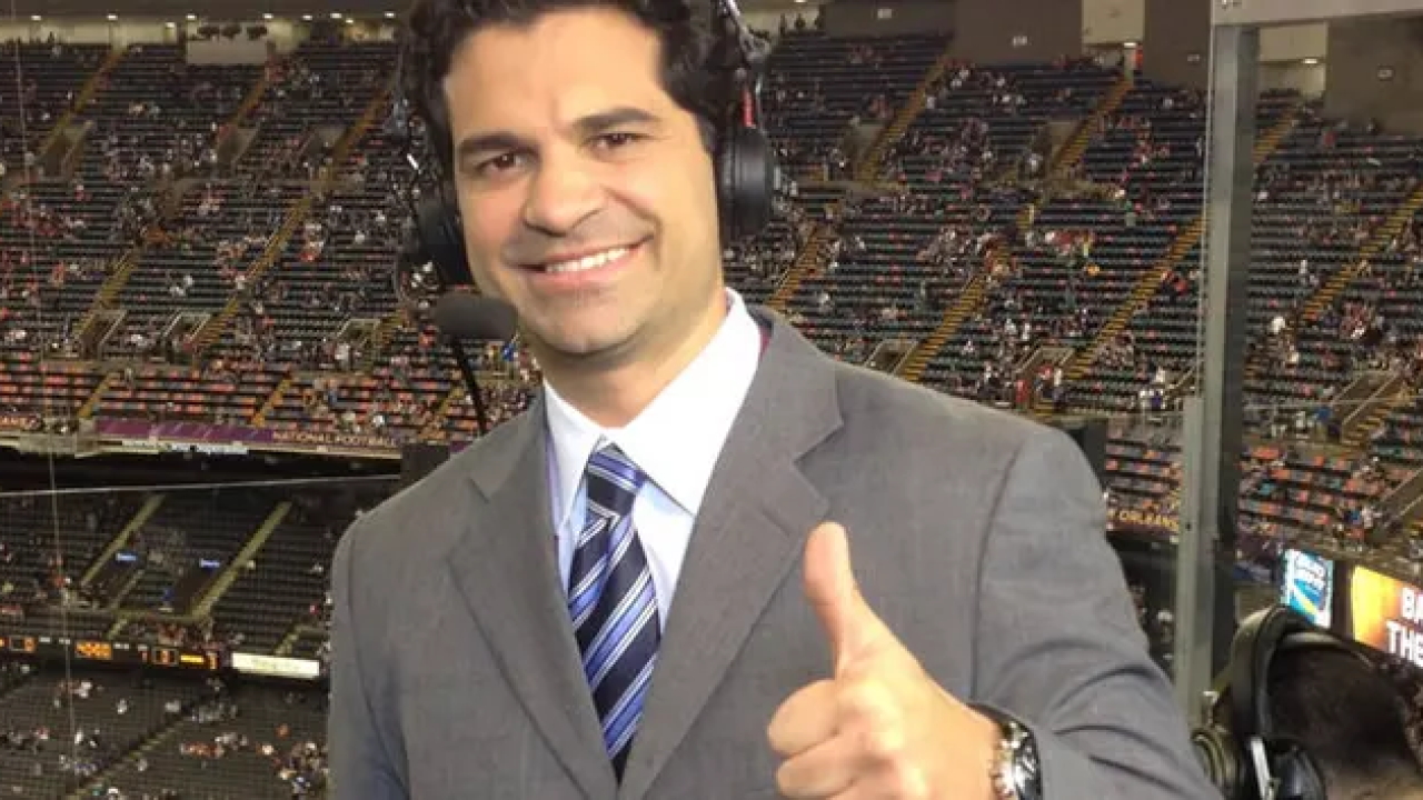 Na imagem, vemos o comentarista da ESPN, Paulo Antunes, em um ambiente esportivo, provavelmente uma cabine de transmissão em um estádio. Ele está usando um terno cinza elegante, com uma camisa branca e uma gravata azul listrada