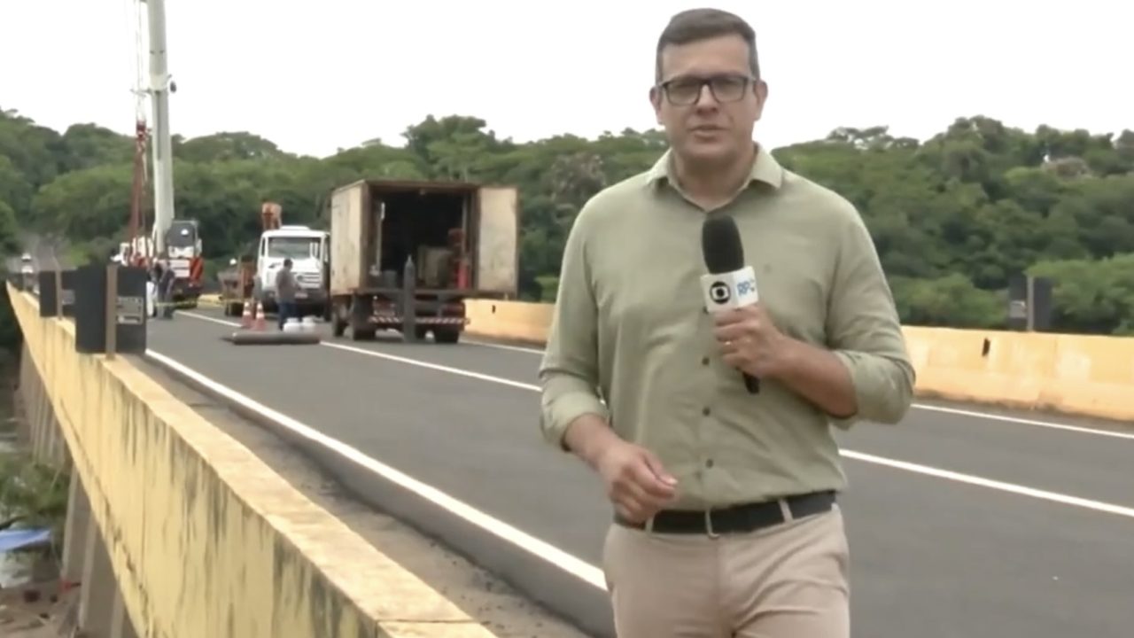 A imagem mostra o jornalista Rodrigo Grando durante uma reportagem em campo para a RPC, afiliada da Globo no Paraná. Ele aparece segurando um microfone com o logo da emissora, vestindo uma camisa social verde clara e calça bege, transmitindo um tom profissional e formal.