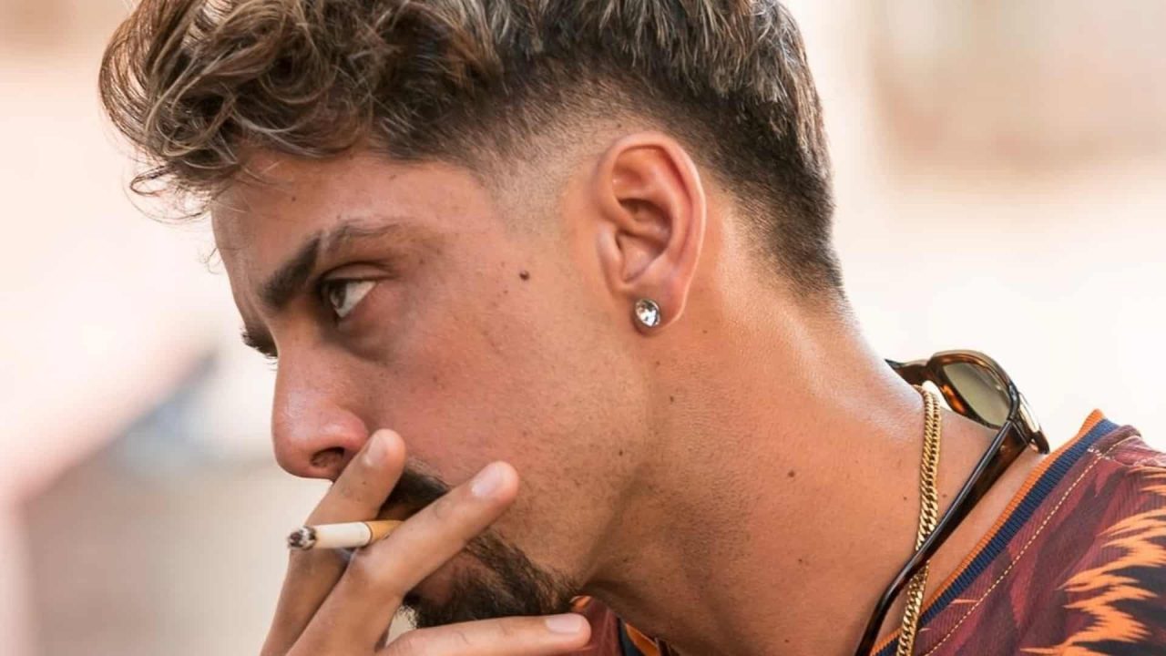 Foto de Rodrigo Simas em Vidas Bandidas fumando cigarro usando brinco e com luzes no cabelo