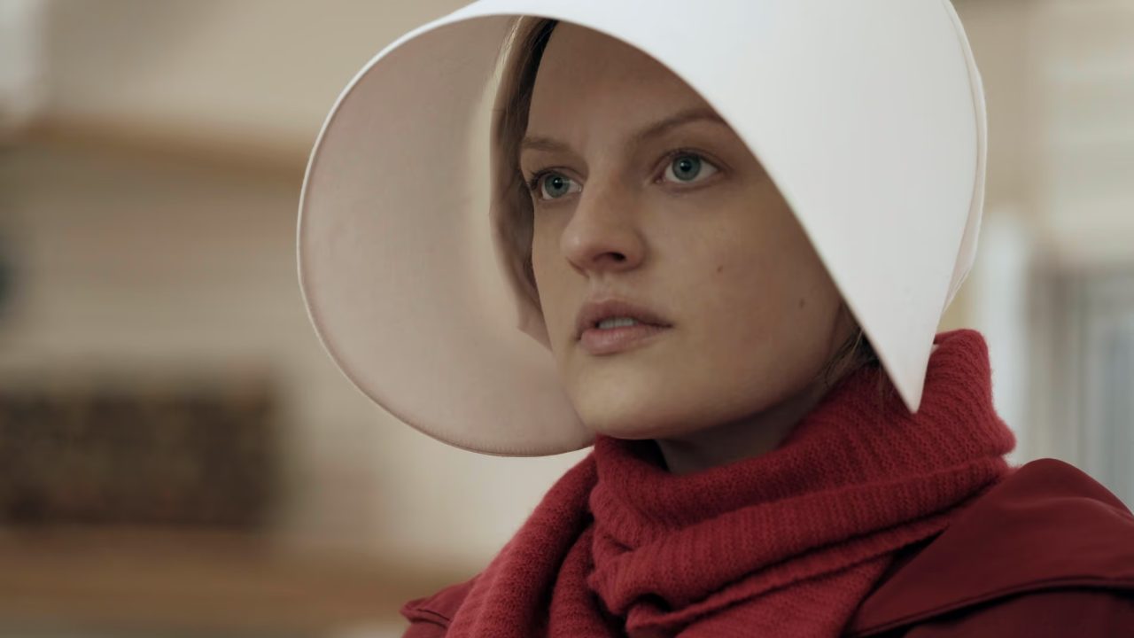 Cena da série The Handmaid’s Tale mostra a protagonista June Osborne, interpretada por Elisabeth Moss, vestida com o característico uniforme vermelho e o icônico chapéu branco das aias. O olhar fixo e sério da personagem transmite tensão e resistência, elementos centrais da trama distópica