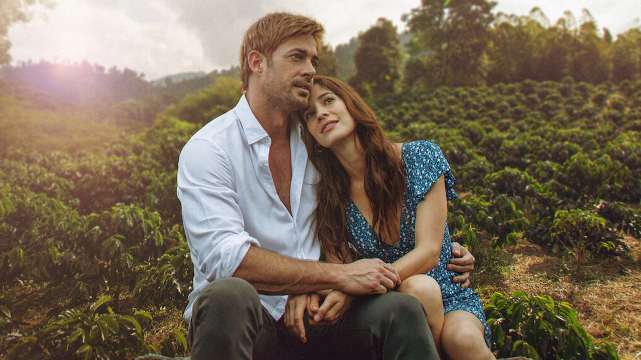 William Levy e Laura Londoño, caracterizados como seus personagens em *Café com Aroma de Mulher*, estão sentados em um campo de café, abraçados e olhando para o horizonte